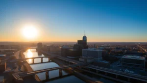 Aerial sunrise view of Rochester New York skyline with Genesee River winding through downtown, morning light casting long shadows across buildings and bridges, clear blue sky