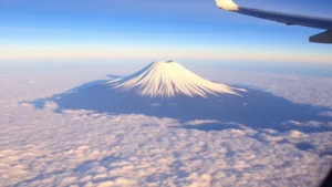 Aerial view of Mount Fuji snow-capped peak at sunrise with clouds below, taken from airplane window showing the dramatic landscape of Japan