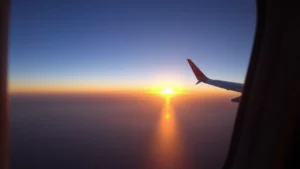 Commercial aircraft window view showing Pacific Ocean horizon at sunset with golden light reflecting on calm water below, taken from cruising altitude