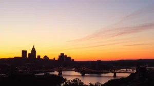 Golden hour sunset over Pittsburgh skyline with Three Rivers and bridges visible, downtown lights beginning to glow, river reflections, urban landscape photography